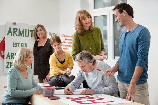 Besprechung Eine Gruppe unterschiedlicher Menschen - Männer, Frauen, Alte und Junge, mit und ohne Behinderung, malt gemeinsam Demo-Plakate.