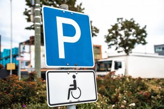 Ein Verkehrsschild weist einen Behindertenparkplatz aus - oben das weiße P auf blauem Grund, darunter ein schwarzes Rollstuhl-Piktogramm auf weißem Grund.
