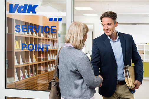 Ein VdK-Mitarbeiter begrüßt eine ältere Frau an der Tür einer VdK-Beratungsstelle. Er hat eine Aktenmappe unter dem Arm. 