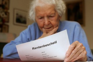 Eine ältere Frau hält ein Dokument in der Hand, auf dem "Rentenbescheid" steht, sie wirkt besorgt oder ratlos.