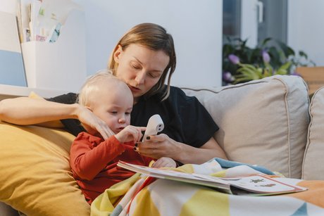 Eine Mutter sitzt mit ihrem Kleinkind auf dem Sofa. Das Kind untersucht ein digitales Fieberthermometer, die Mutter erklärt ihm etwas.