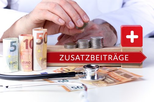 Zusatzbeiträge der Krankenkassen Hände legen Münzen auf gestapelte Euro-Münzen und Geldscheine; daneben ein roter Pfeil mit der Aufschrift „Zusatzbeiträge“, ein roter Würfel mit weißem Kreuz und ein Stethoskop – Symbolbild für steigende Zusatzbeiträge im Gesundheitswesen.