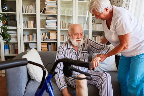 Ein älterer Mann im gestreiften Pyjama sitzt auf einem Sofa. Eine ältere Frau hält ihn am Arm und hilft ihm aufzustehen. Im Vordergrund steht ein Rollator. 