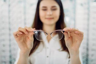 Eine junge Frau hält mit beiden Händen eine Brille hoch. Im Hintergrund sieht man Regale mit vielen Brillenfassungen.