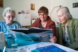 Eine Frau liest zwei Seniorinnen in einem Pflegeheim aus einem Bildband vor. 