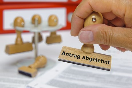 Eine Hand stempelt ein Dokument mit dem Stempel mit der Aufschrift "Antrag abgelehnt". Im Hintergrund ein Gestell mit drei weiteren Stempeln.