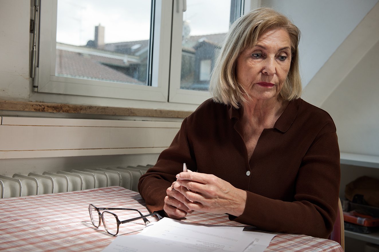 Eine &auml;ltere Frau sitzt an einem K&uuml;chentisch, sie sieht besorgt und niedergeschlagen aus. Auf dem Tisch vor ihr liegt ein Brief, daneben ihre Lesebrille. 