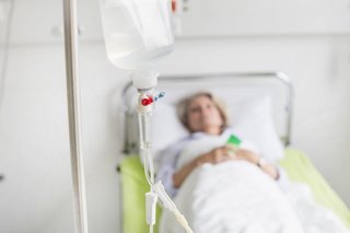 Eine Frau liegt in einem Krankenhausbett, im Vordergrund ein Tropf 