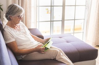 Eine ältere Frau sitzt auf einem Sofa, liest in einem Buch.