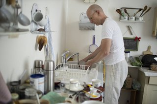 Ein Mann in weißer Kleidung steht in einer Küche und spült mit der Hand Geschirr.