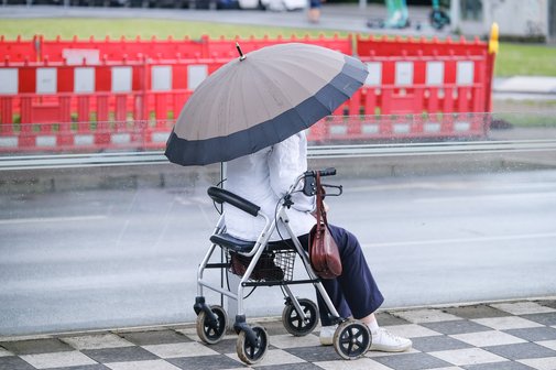 Eine Frau mit aufgespanntem Regenschirm sitzt auf ihrem Rollator auf einem Platz. Im Hintergrund ist ein Baustellenzaun zu sehen.
