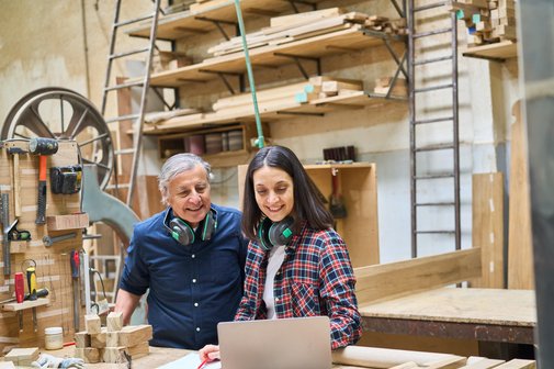 Ein älterer Mann und eine Frau in einer Schreinerei, sie beugen sich gemeinsam über ein Notebook, im Hintergrund Holz, Werkzeuge und Maschinen