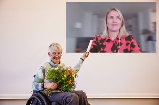 Dr. Sigrid Arnade hält den DBR-Staffelstab in die Höhe, wo auf einer Leinwand Verena Bentele digital zugeschaltet ist, und übergibt ihr damit symbolisch den DBR-Vorsitz für das nächste Jahr.