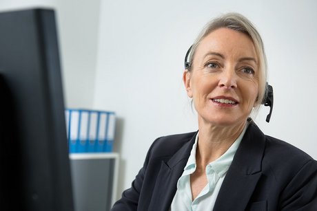 Eine Frau am Computer, sie telefoniert über ein Headset. Im Hintergrund sieht man Aktenordner.