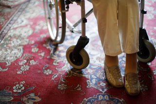 Beine einer Seniorin im Rollstuhl, er steht auf einem rot gemusterten Perserteppich 