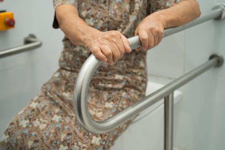 Eine &auml;ltere Frau stzt auf einem geschlossenen Toilettendeckel, sie h&auml;lt sich mit beiden H&auml;nden an einem Haltegriff fest, der neben der Toilette angebracht ist.