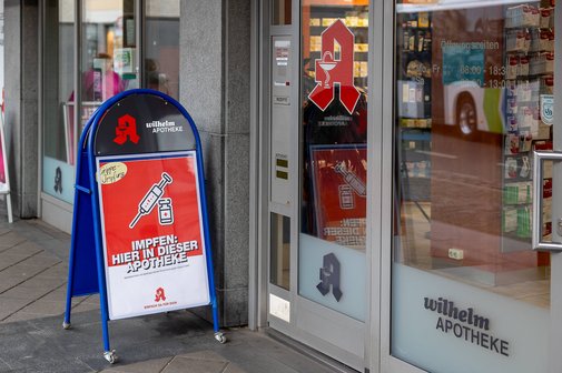 Eine Apotheke mit einem Aufsteller vor dem Eingang, der auf die Möglichkeit einer Grippeschutzimpfung hinweist. Das Schild trägt die Aufschrift Impfen: Hier in dieser Apotheke und zeigt ein Symbol einer Spritze und eines Impfstoff-Fläschchens.