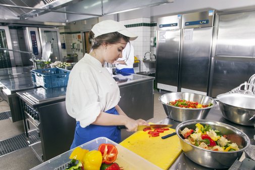 Arbeiten zum Mindestlohn Eine junge Frau in Arbeitskleidung arbeitet in einer Großküche, sie schneidet Tomaten