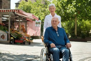 Älteres Paar draußen unterwegs, er sitzt im Rollstuhl, sie schiebt ihn