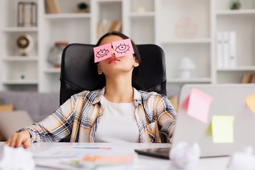 Eine junge Frau sitzt am Schreibtisch in einem Büro, sie hat den Kopf zurückgelegt. Auf ihren geschlossenen Augen liegen Post-it-Zettel mit aufgemalten offenen Augen.