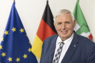 Das Foto zeigt Karl-Josef Laumann, im Hintergrund sind drei Flaggen zu sehen: Die Europa-Flagge, die Deutschlandflagge und die Flagge von Nordrhein-Westfalen