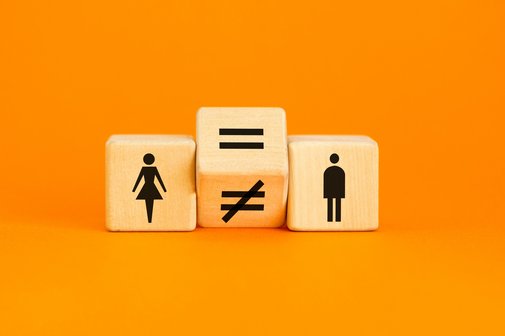 Drei Würfel liegen nebeneinander vor orangenem Hintergrund. Einer zeigt ein Piktogramm einer Frau, einer das eines Mannes. Dazwischen liegt ein gekippter Würfel auf der Kante, der ein Gleichheits- und ein Ungleichheitszeichen anzeigt.
