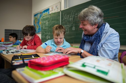 Blick in eine Schulklasse, eine Inklusionshelferin kümmert sich um einen Jungen mit Trisomie 21. 