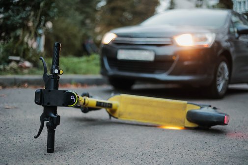 Ein gelber E-Scooter liegt umgestürtzt auf der Straße. Ein Auto, dessen Scheinwerfer an sind, steht weniger Meter entfernt. 