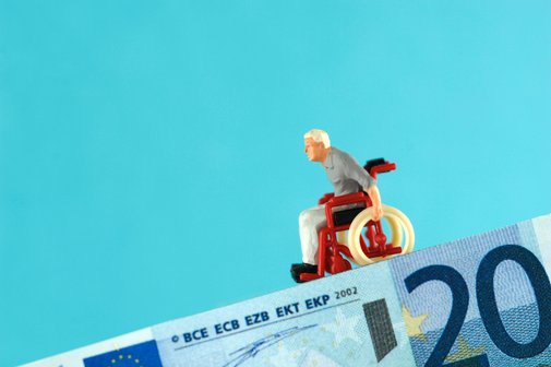Symbolbild: Ein 20-Euro-Schein bildet eine abschüssige Rampe, auf der eine Mini-Figur eines alten Mannes im Rollstuhl bergab rollt