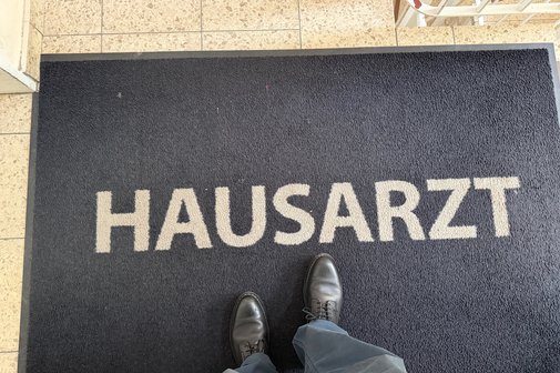 Jemand steht auf einer Fußmatte, auf der "Hausarzt" in Blockbuchstaben geschrieben steht