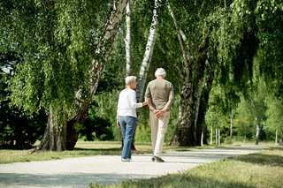 Ein älterer Mann und eine ältere Frau gehen gemeinsam auf einem Weg durch einen Wald oder Park spazieren. 