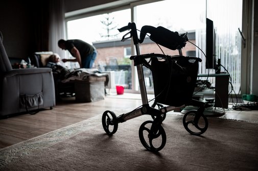 Blick in ein dunkles Wohnzimmer. Im Vordergrund steht ein Rollator. Im Hintergrund sieht man eine sitzende Person, die von einer anderen Person, die sich über sie beugt, gefüttert wird. Symbolfoto für Angehörigenpflege