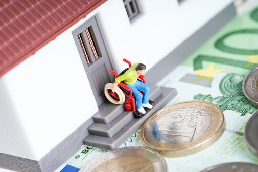 Ein Spielzeughaus, auf der Türschwelle sind Stufen. Ein Figürchen im Rollstuhl steht an der Schwelle, schaut nach unten. Vor dem Spielzeughaus liegen Geldscheine und -münzen.
