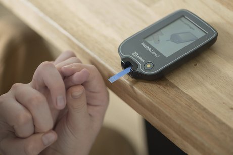 Ein Blutzucker-Messgerät mit Teststreifen liegt auf einem Holztisch, eine Kinderhand hält einen Finger fest und führt ihn zum Messgerät.