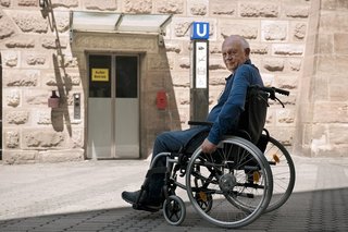 Ein Senior im Rollstuhl steht am unzugänglichen Eingang einer U-Bahn-Station. Dort ist ein Aufzug mit dem Hinweis "Außer Betrieb". 