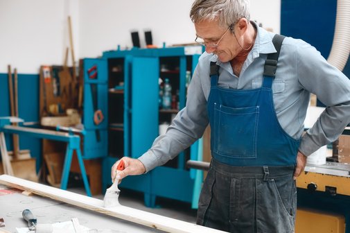 Ein älterer Handwerker mit grauem Haar und Brille streicht eine Leiste mit weißer Farbe, hat eine Hand in den Rücken gestützt, trägt einen Blaumann