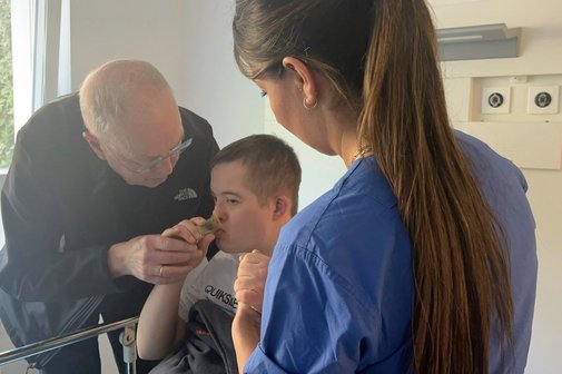 Ein Junge mit Down-Syndrom wird von einer jungen Frau im Arztkittel untersucht, der Vater steht neben dem Kind und beruhigt es.