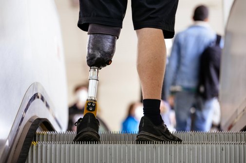 Ein Mann mit einer Beinprothese fährt Rolltreppe. Man sieht nur seine Füße und Waden, an einem Bein hat er eine Unterschenkelprothese.
