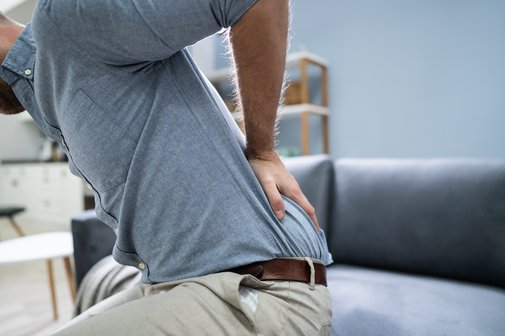 Ein Mann steht vom Sofa auf, h&auml;lt sich mit einer Hand den unteren R&uuml;cken als Symbol f&uuml;r R&uuml;ckenschmerzen