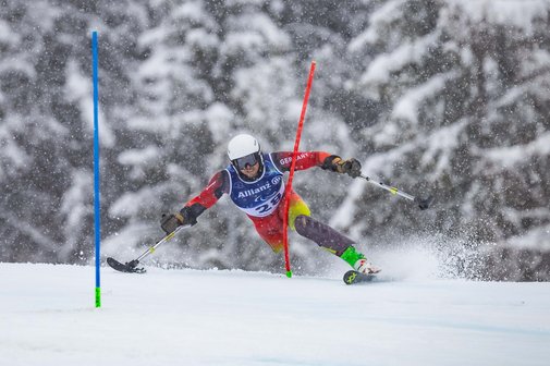  VdK-Mitglied Christoph Glötzner fährt zwischen den Slalomstangen durch. Mit einem Bein, dem linken, einem Ski und zwei Armstützen, die unten kleine, gerundete Flächen haben und so auf dem Schnee gleiten, meistert er den Kurs. Man sieht auf dem Foto, dass es schneit und eingeschränkte Sicht ist.