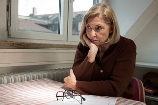 Eine ältere Frau sitzt an einem Küchentisch, sie sieht besorgt und niedergeschlagen aus. Auf dem Tisch vor ihr liegt ein Brief, daneben ihre Lesebrille. Sie stützt den Kopf in die Hand.