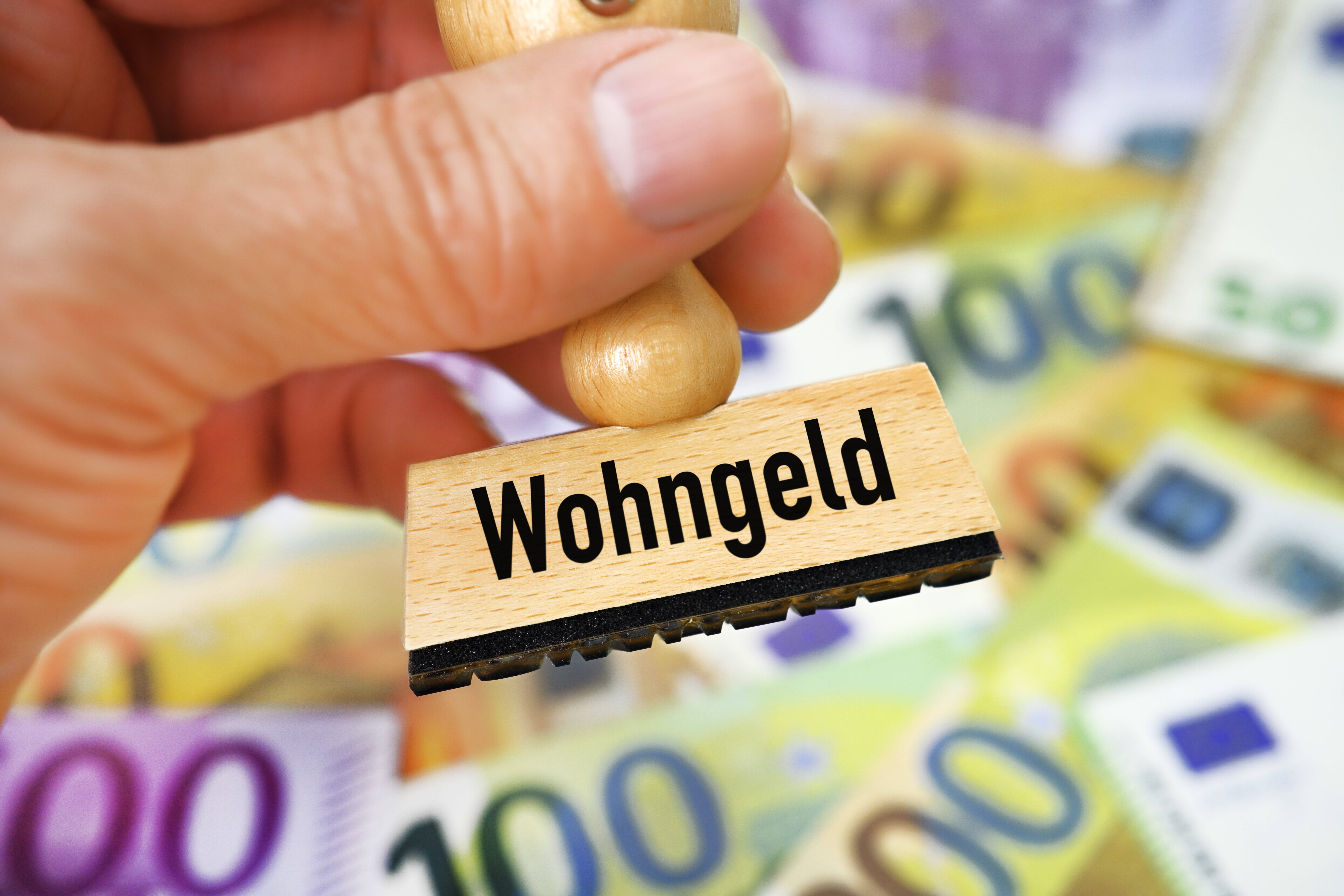 Wohngeld Eine Hand hält einen Stempel mit der Aufschrift Wohngeld, im Hintergrund viele Geldscheine