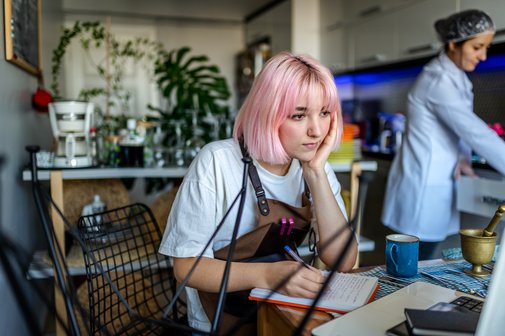 Studentin arbeitet in einem Café als Minijob, sie steht an der Theke und blickt, auf ihren Ellbogen geschützt, auf die Kasse, rechts von ihr im Hintergrund steht eine Kollegin.