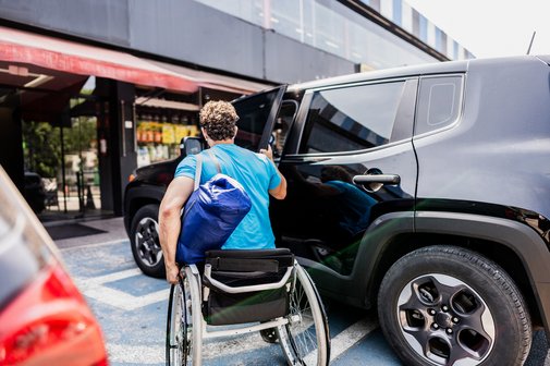 Mann im Rollstuhl öffnet auf einem Parkplatz die Tür zu seinem Auto, um einzusteigen.