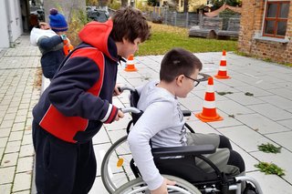 Auf dem Fotosieht man zwei junge Menschen beim Parcours. Einer der beiden Jungs sitzt im Rollstuhl, der andere schiebt den Rollstuhl.