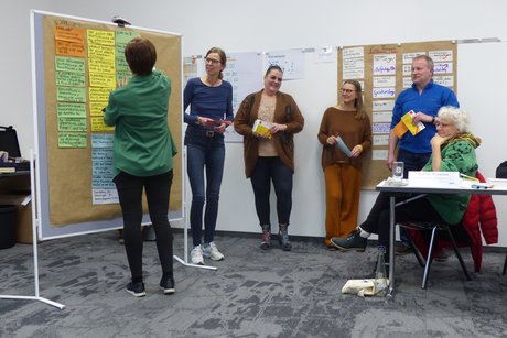 Das Foto zeigt Teilnehmende der Schwerbehindertenschulung während eines Workshops. Eine Frau steht an einer Pindwan mit vielen bunten beschrifteten Zetteln. die anderen Teilnehmer betrachten die Pinwand. 