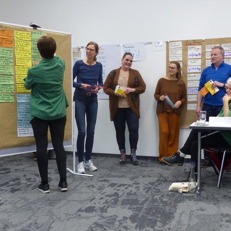 Das Foto zeigt Teilnehmende der Schwerbehindertenschulung während eines Workshops. Eine Frau steht an einer Pindwan mit vielen bunten beschrifteten Zetteln. die anderen Teilnehmer betrachten die Pinwand. 