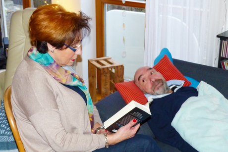 Auf dem Foto sieht man Margit Heßdörfer und Edgar Nestmeier. Sie sitzt auf einem Stuhl und hält ein Buch in ihrer Hand. Er liegt auf dem Sofa.