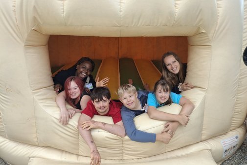 Zwei Freizeitbegleiterinnen und vier Kinder, zwei M&auml;dchen und zwei Jungen, schauen aus dem Eingang zu einer H&uuml;pfburg. Sie lachen. 