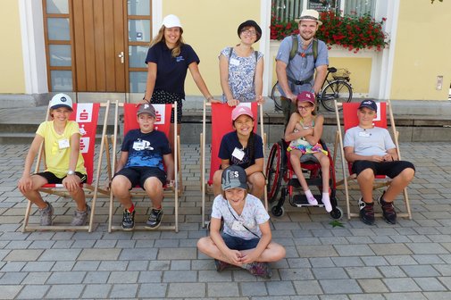 Das Foto zeigt die Teilnehmend und ihre Betreuerinnen und Betreuer der inklusiven Ferienfreizeit vor einem Haus. Die Teilnehmenden sitzen gl&uuml;cklich in Liegest&uuml;hlen.